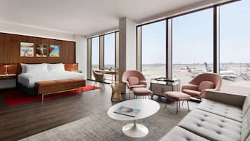 A modern hotel room with a large bed, two armchairs, a sofa, a small round table, and a view of an airport.