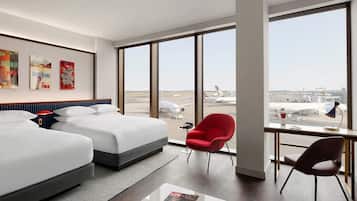 A hotel room with two beds, a desk, and a large window with an airport view.