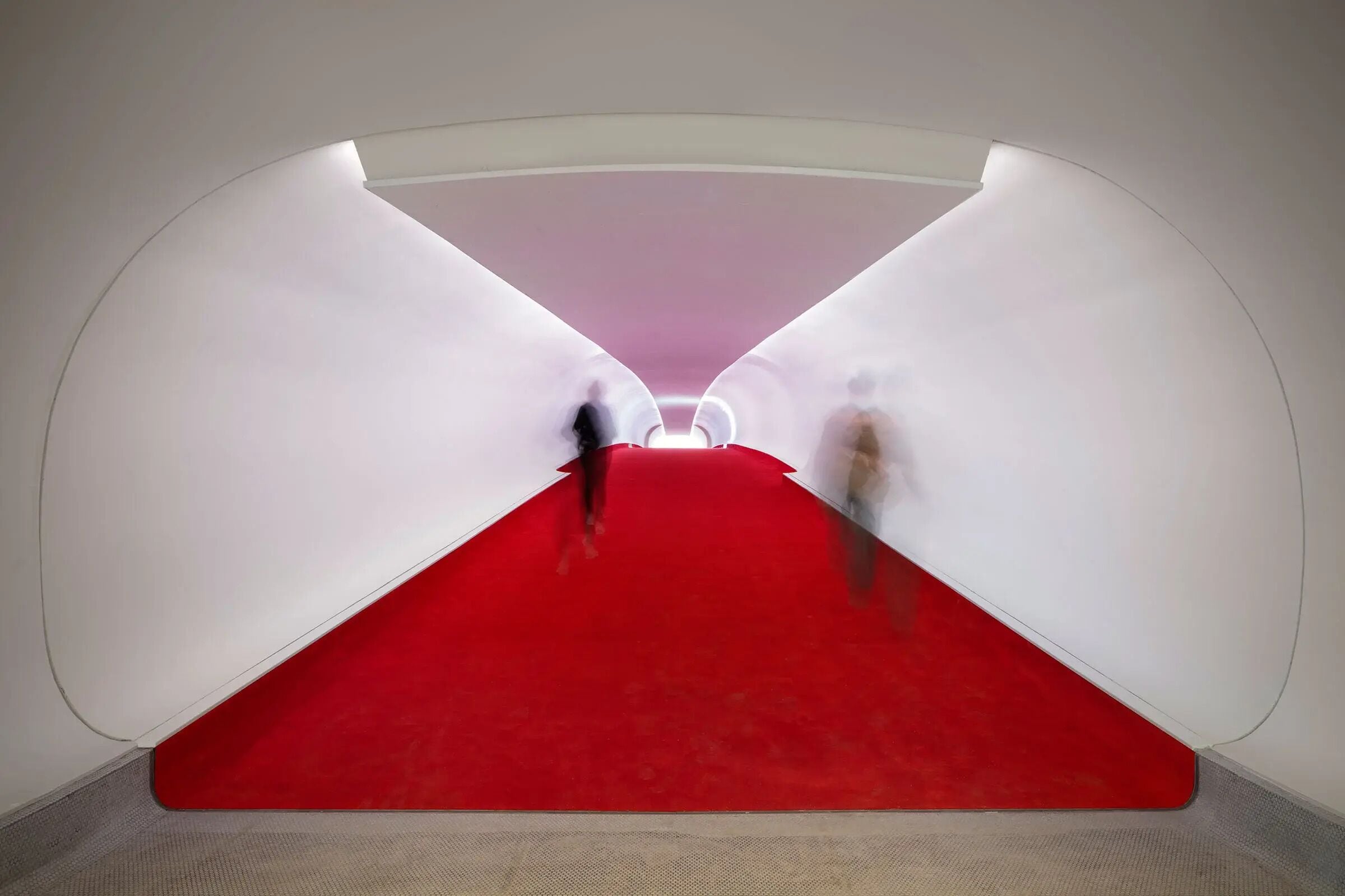 Une pièce dont le sol est recouvert d’une moquette rouge, et qui mène à un mur blanc orné d’un luminaire circulaire.