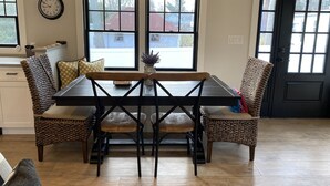 A dining area with a wooden table and chairs, two windows, and a clock on the wall. - Charming Brand New Beach House (Mattituck)