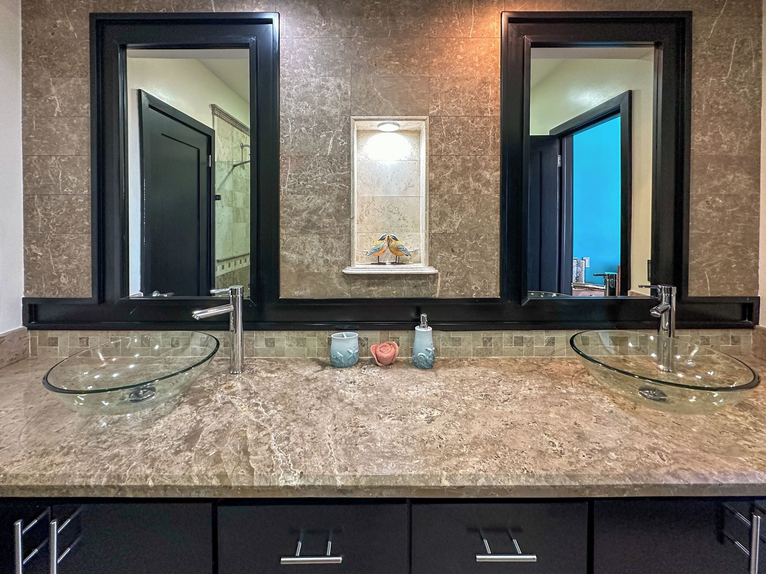 A bathroom with two large mirrors, a glass sink, and a small shelf with a decorative item.