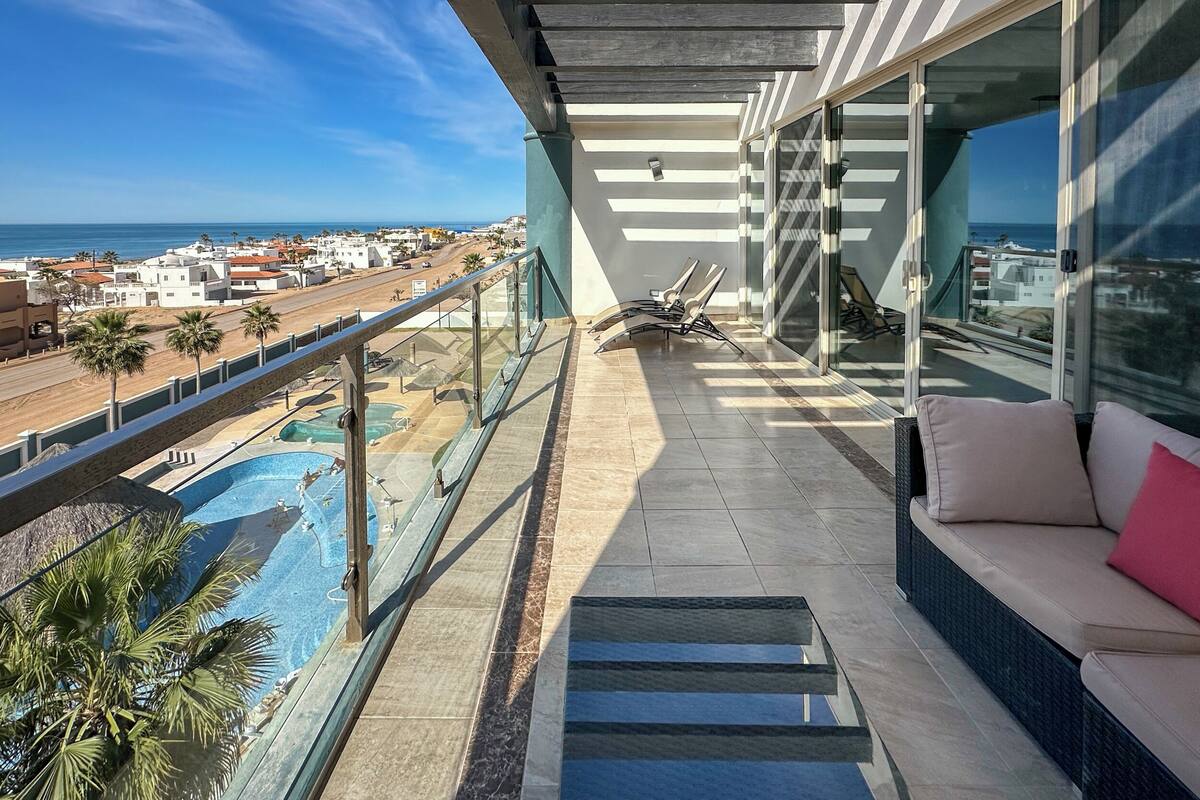 Un balcón con vista al mar, una piscina y una zona de estar con cojines.