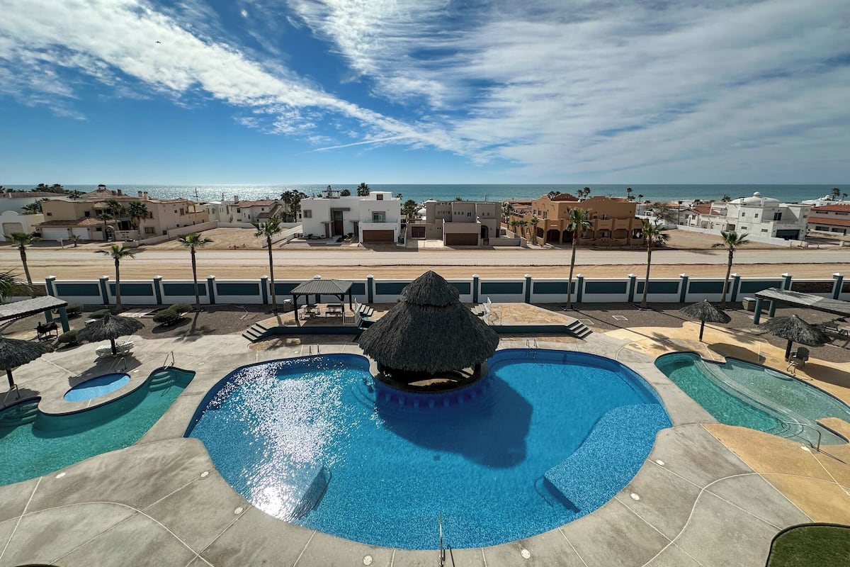Zona de piscina con una fuente central, rodeada de sillas de descanso y sombrillas, y vista a la playa y al océano.