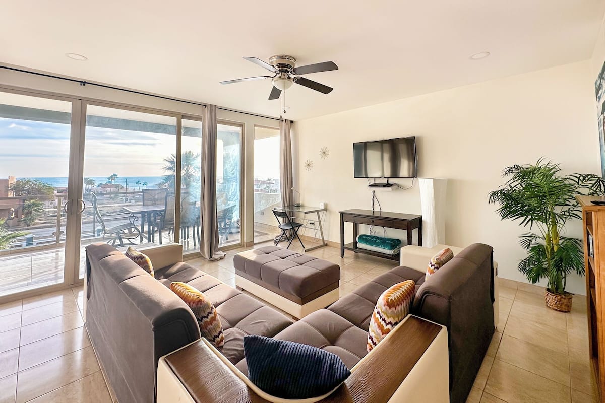 Amplia sala de estar con un amplio sofá, ventilador de techo, televisor de pantalla plana y vistas a la ciudad a través de amplias ventanas.