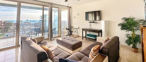 A spacious living room with a large sofa set, a ceiling fan, a flat-screen TV, and a view of the city through large windows.