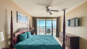 Ein Bett mit Baldachin, mit einem türkisfarbenen Überwurf, ein Deckenventilator, ein Fernseher an der Wand und ein Balkon mit Meerblick.