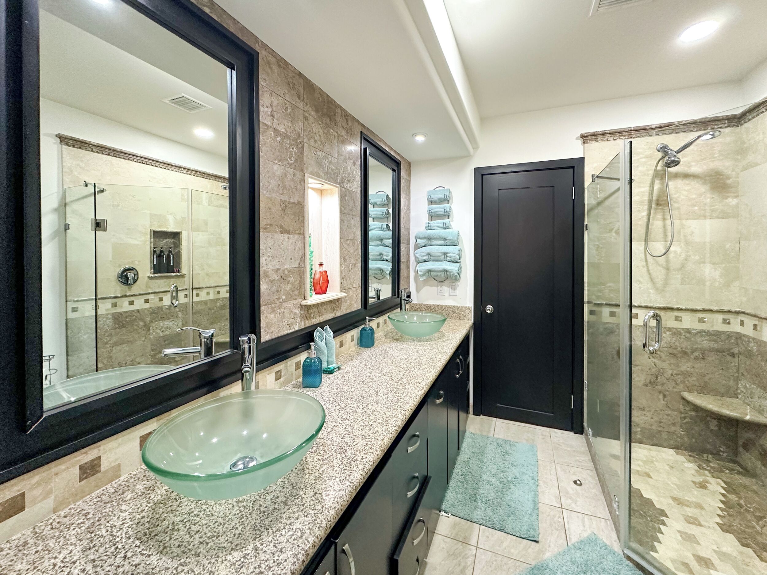 A modern bathroom with a glass bowl sink, a large mirror, a walk-in shower, and a dark door.