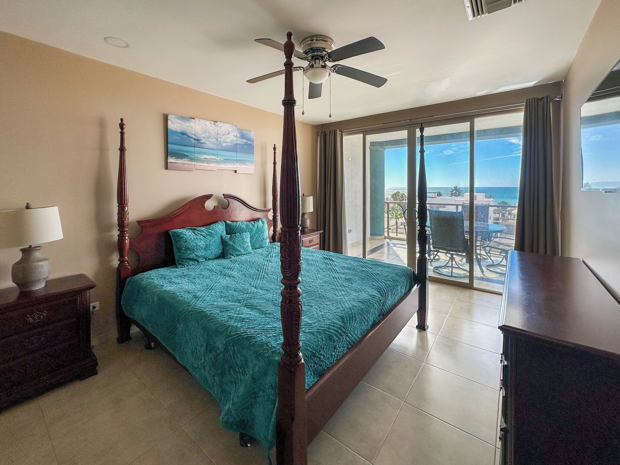 Un dormitorio con cama con dosel, ventilador de techo, ventana grande con vistas y mesita de noche con lámpara.