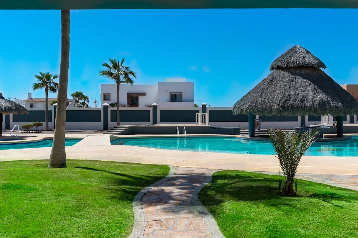 Una piscina con una estructura de techo de paja, palmeras y edificios modernos al fondo.