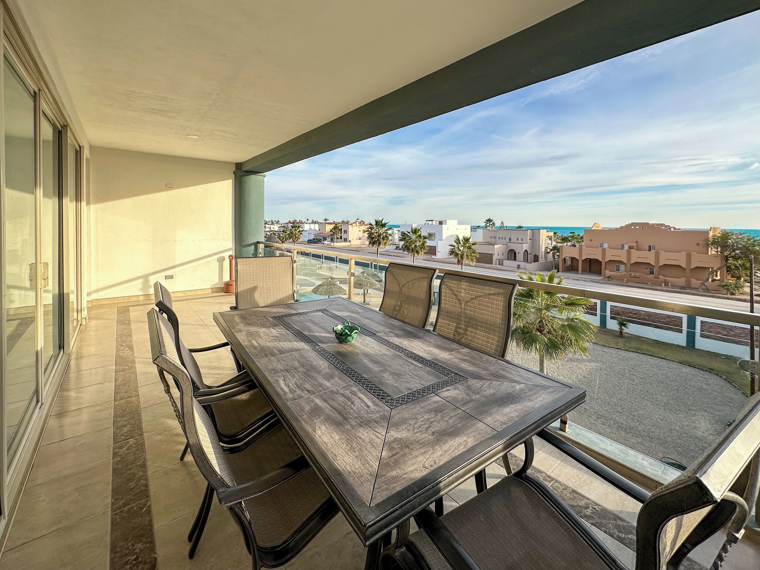 A balcony with a table and chairs, overlooking a coastal area with buildings and palm trees.