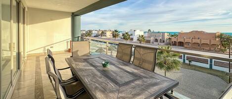 A balcony with a table and chairs, overlooking a coastal area with buildings and palm trees.