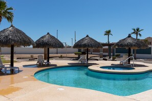 Zwembad met ligbedden en rieten parasols, omringd door palmbomen en een heldere lucht.