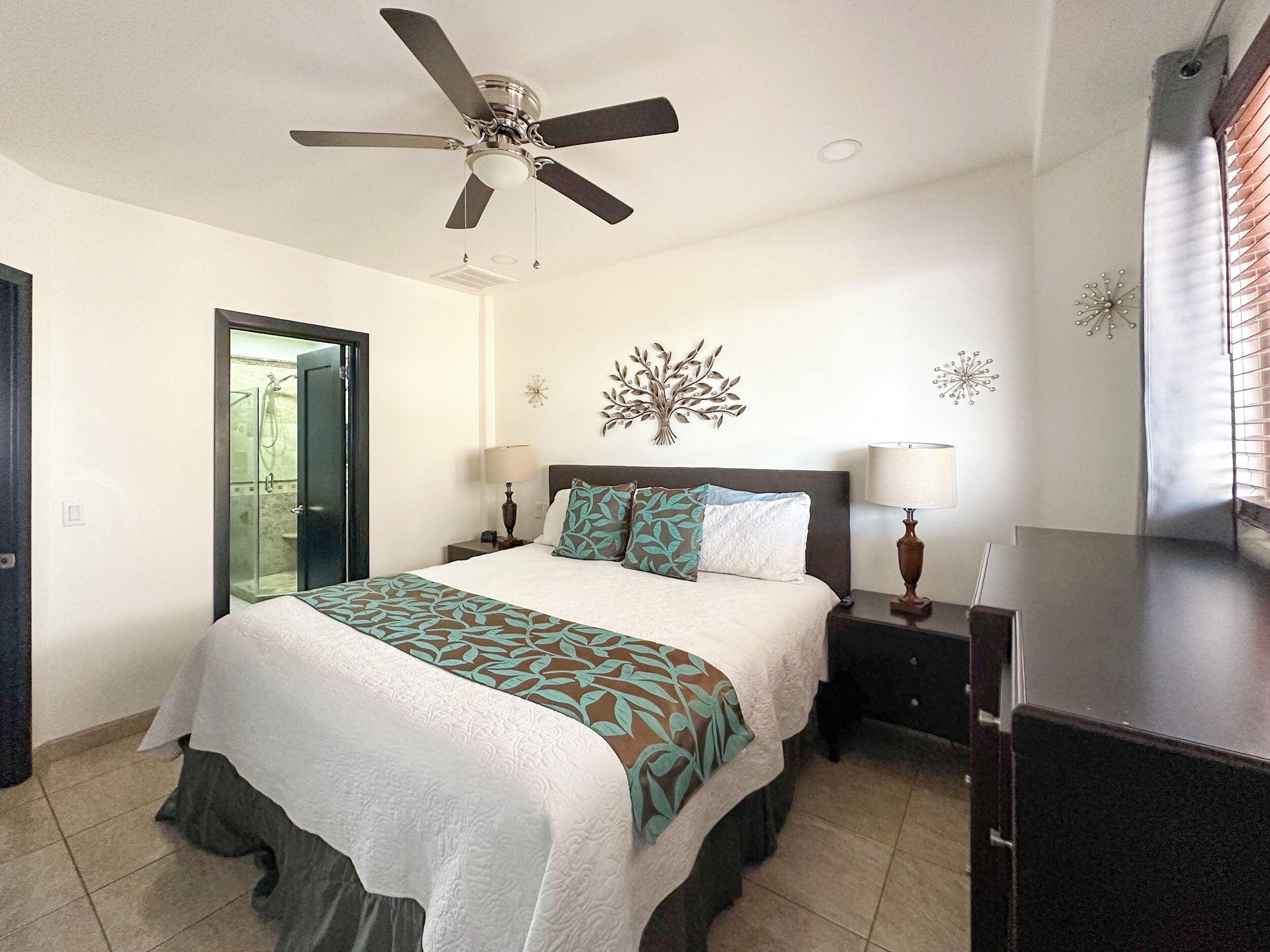 A bedroom with a bed, a ceiling fan, a nightstand, a dresser, and a door leading to a bathroom.