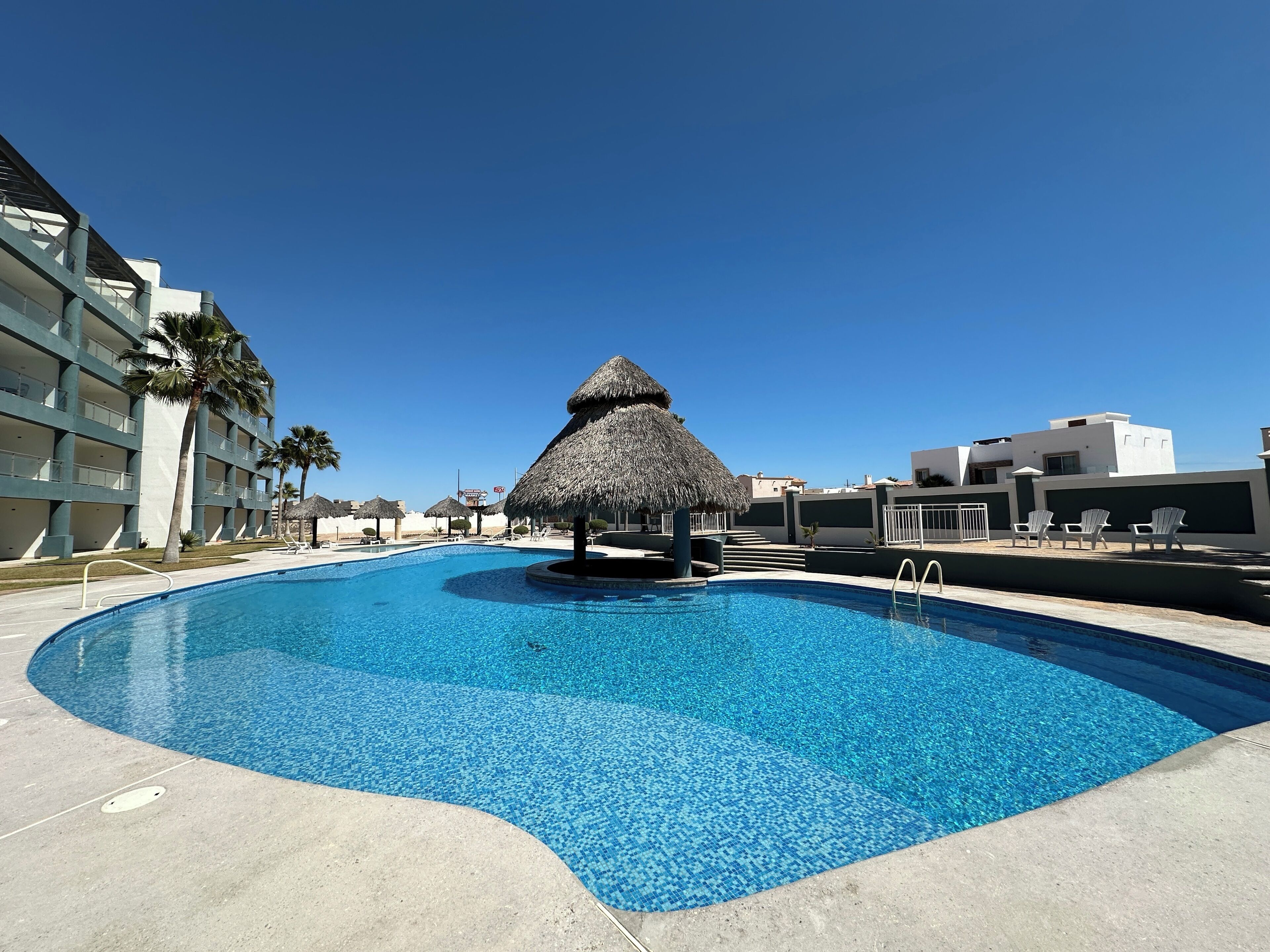 Una piscina con una estructura de techo de paja, rodeada por un edificio de varios pisos con balcones y palmeras.