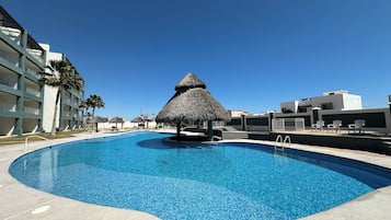 Una piscina con una estructura de techo de paja, rodeada por un edificio de varios pisos con balcones y palmeras.