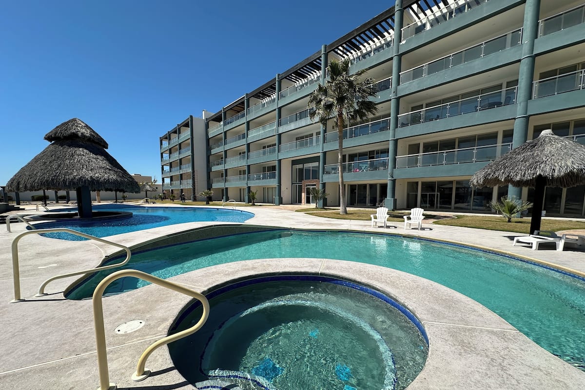 Un hotel con piscina, sombrillas de paja y un edificio de varios pisos con balcones.