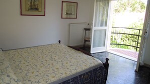 Ein Schlafzimmer mit einem Bett, zwei gerahmten Bildern an der Wand, einem Sessel und einem Balkon mit Blick auf Grünflächen.
