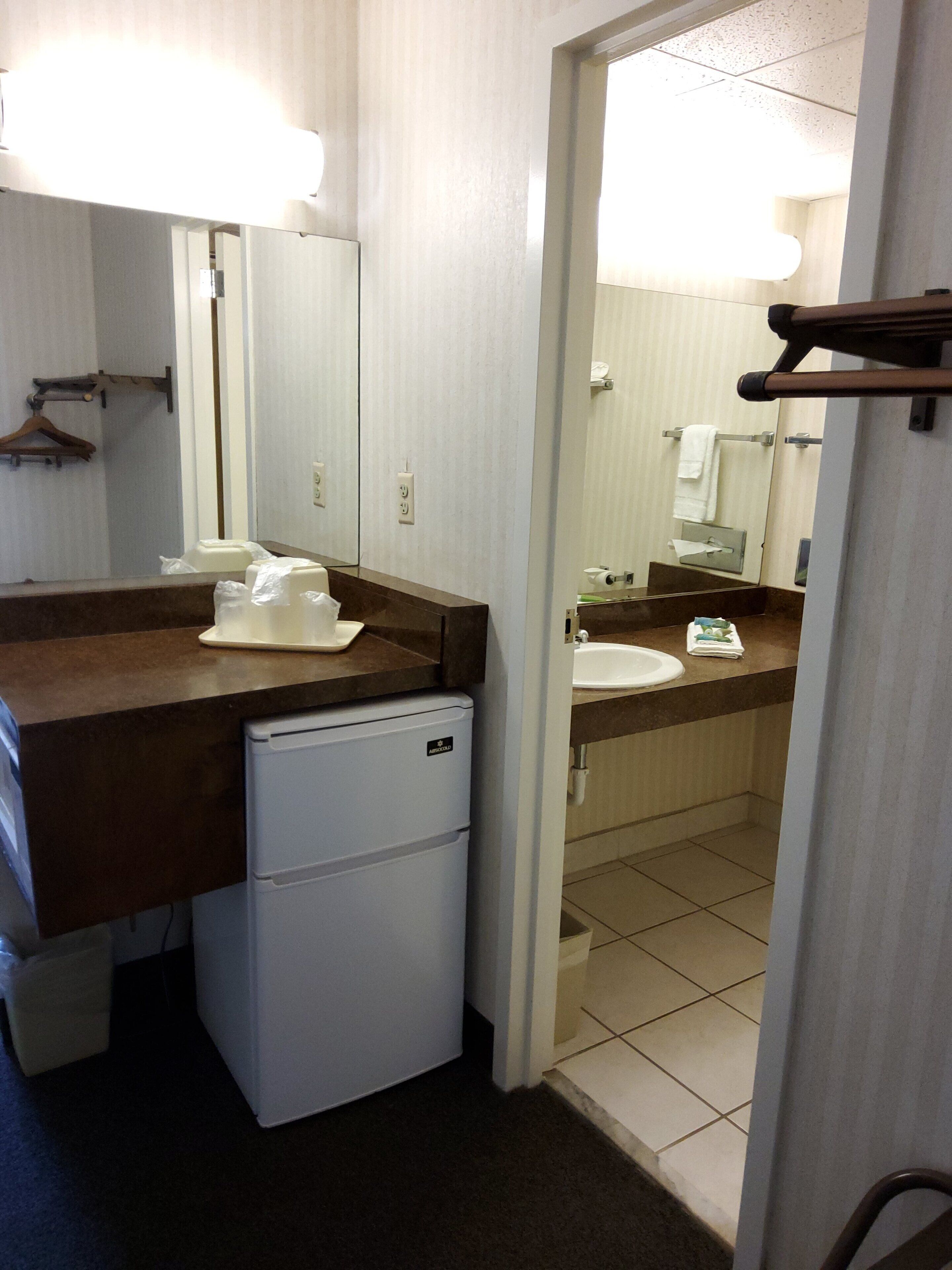 Een kleine hotelbadkamer met een koelkast, wastafel en spiegel.