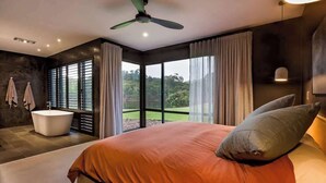 A bedroom with a large bed, a ceiling fan, a bathtub, and a view of greenery through the windows. - Artisan Gunyulgup—Luxe Lakeside Wellness Hideaway (Yallingup)