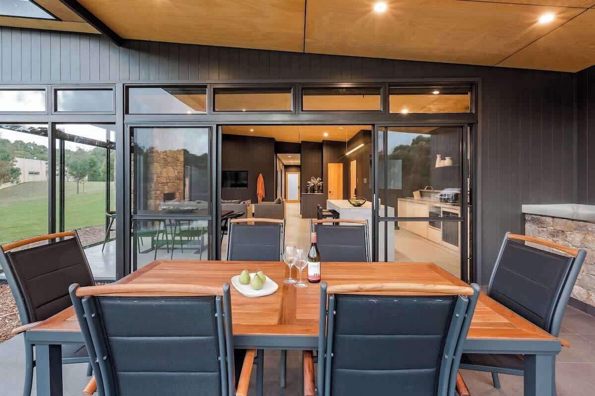 An outdoor dining area with a wooden table and chairs, a stone wall, and a view of the interior.