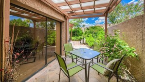 Un patio coperto con una porta vetrata che si apre su un giardino, un tavolo con sedie e un pergolato sovrastante.