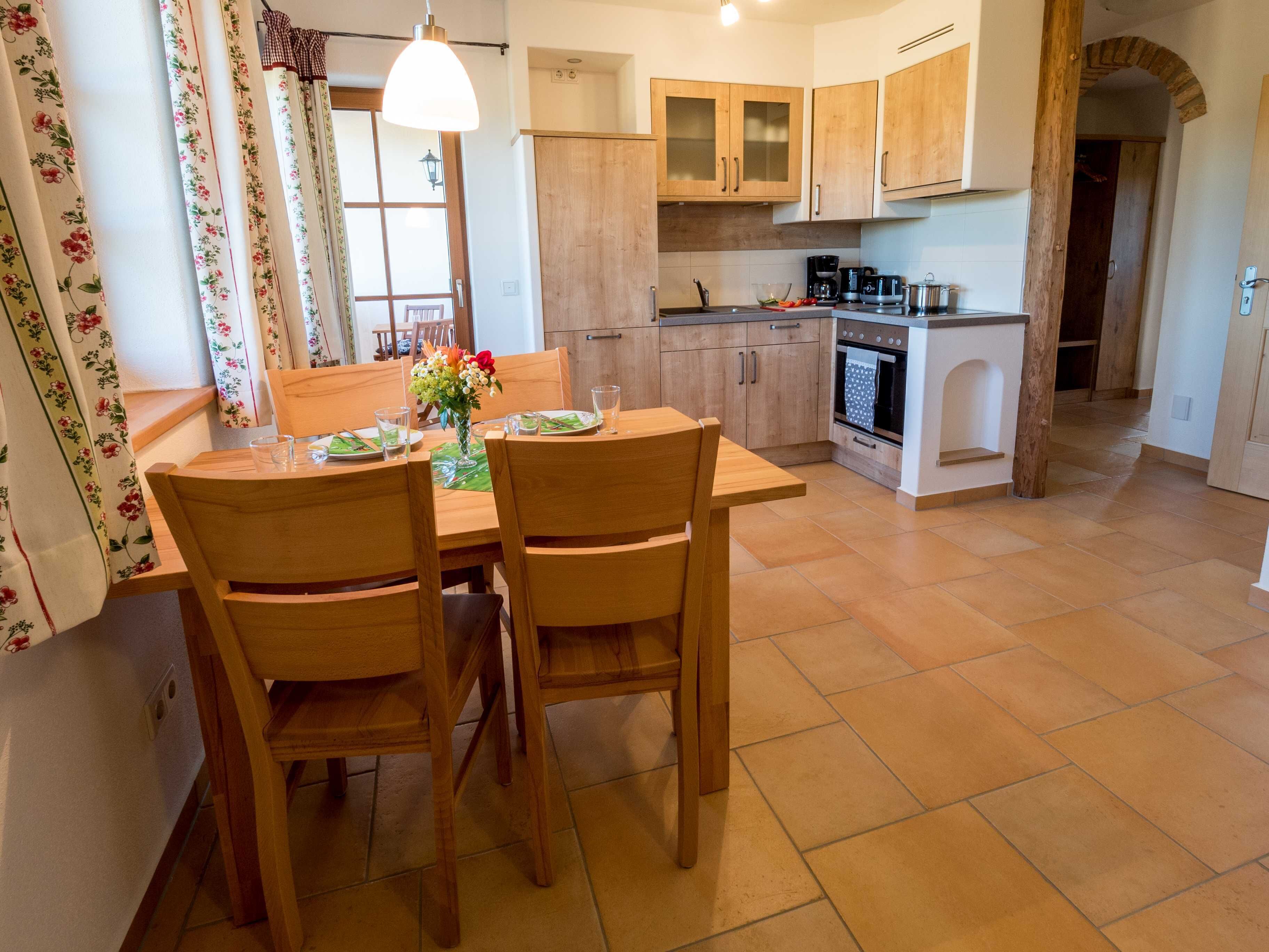 Een eethoek met een houten tafel en stoelen, een bloevas en uitzicht op de keuken met houten kasten en een fornuis.