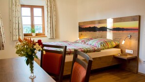 Quarto com cabeceira de madeira, mesa de cabeceira com abajur, janela com cortinas e um arranjo floral sobre uma mesa.