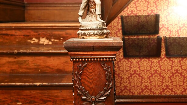 Eine Statue auf einem hölzernen Sockel mit aufwendigen Schnitzereien, vor einer gemusterten Wand und einer Holztreppe.