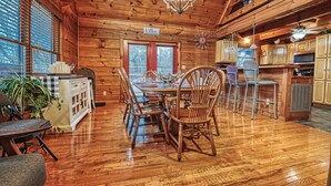 A wooden cabin interior with a dining table, chairs, a kitchen area, and a large window with blinds.
