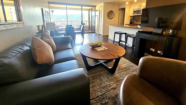 A living room with a sofa, coffee table, and chairs, a kitchen area with a TV, and large windows offering a view. - Great Location In Wakiki, Walking Distance To Beach, Shopping, Dining! (Honolulu)