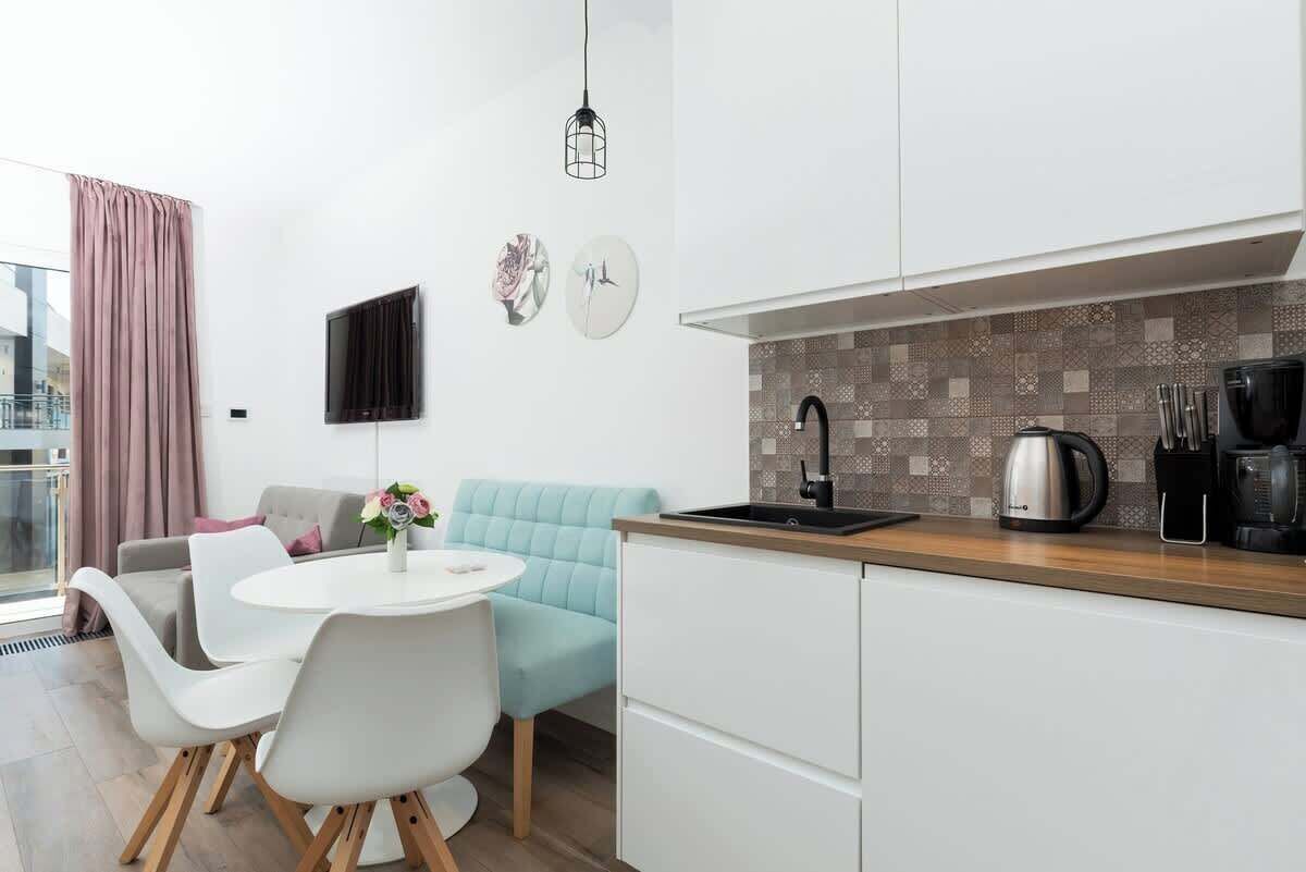 Een moderne keuken met witte kastjes, een betegelde spatwand en een koffiezetapparaat. Er is een eethoek met een ronde tafel en witte stoelen.