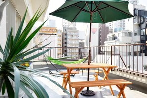 Een balkon met een houten tafel en bankje, een groene parasol en een hangmat.