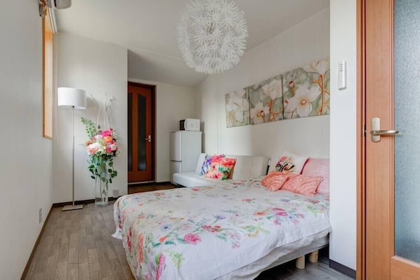 A bedroom with a floral bedspread, a standing lamp, a painting on the wall, and a wooden door.