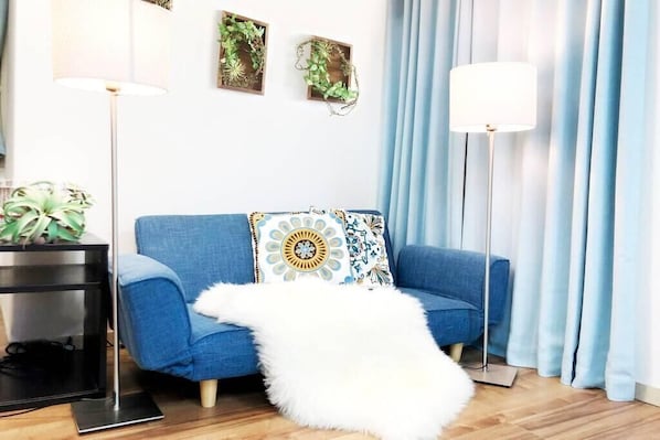 Una habitación con un sofá azul, un plaid blanco mullido, una lámpara de pie, una mesita de noche con una planta y decoración en la pared.