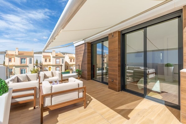 Un espace de vie extérieur moderne doté d’une terrasse en bois, d’un coin salon abrité et d’une porte vitrée donnant accès à une pièce.