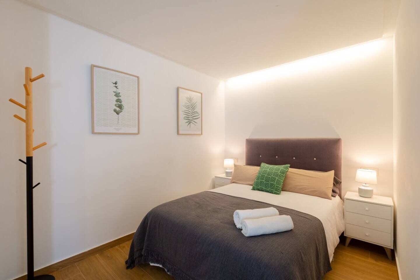 A bedroom with a bed, two bedside tables, a coat rack, and two framed botanical prints on the wall.