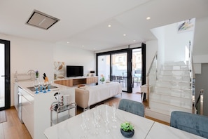 Un appartement moderne comprenant une cuisine, un coin repas et un salon. Il est doté d’un escalier, d’une télévision et d’un balcon.