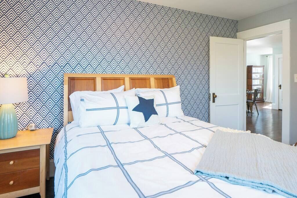 Ein Schlafzimmer mit gemustertem Wandteppich, einem Bett mit einem sternförmigen Kissen, einem Nachttisch mit Lampe und einer Tür zu einem weiteren Raum.