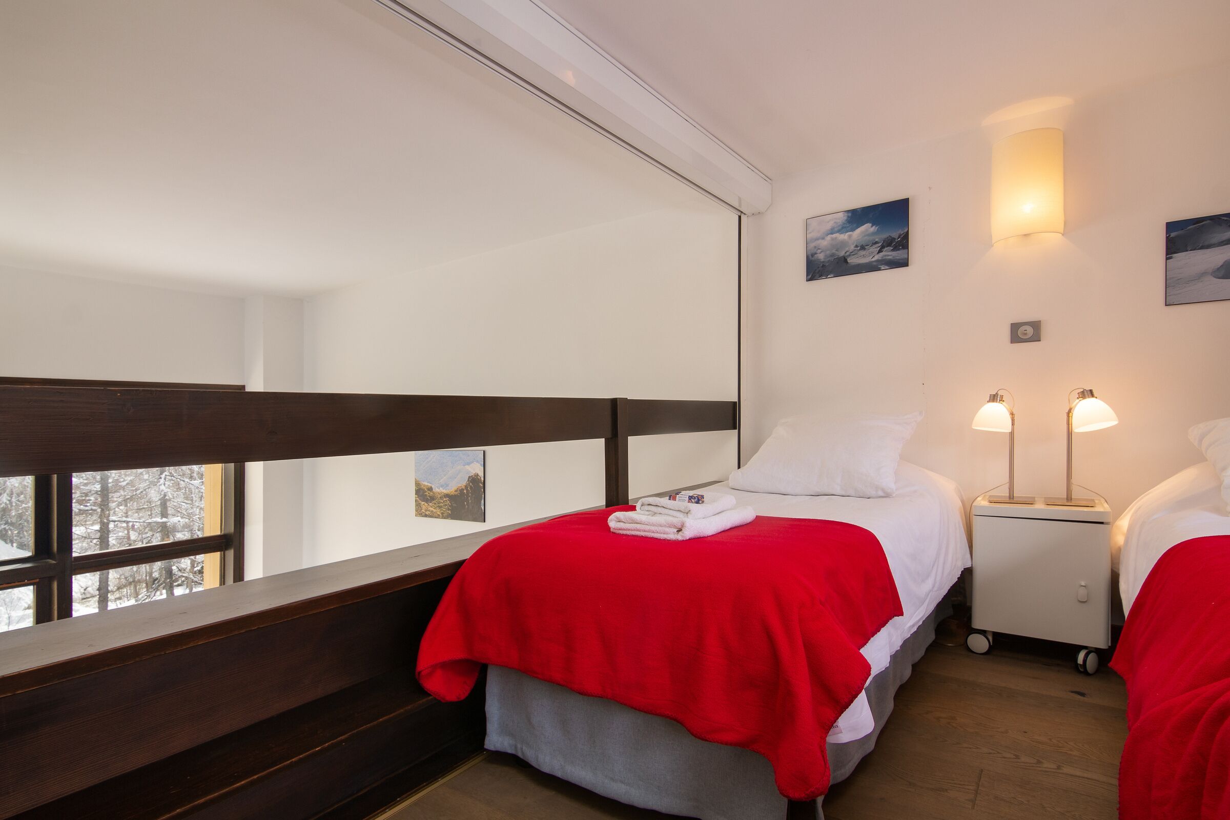 Une chambre d’hôtel avec une grande fenêtre, un lit recouvert d’une couverture rouge, deux tables de chevet avec des lampes et un petit chariot.