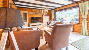 Un interno accogliente di una baita con soffitto in legno, zona pranzo, televisione e vista sull'esterno.