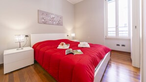A bedroom with a red bedspread, white nightstands, a bedside lamp, a decorative wall piece, and a window with blinds. - Appartamento Il Golfo: Lake View in Laveno-Mombello, Laveno Mombello, Italy (Laveno Mombello)