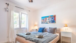 A bedroom with a wooden bed, white curtains, a window, a wall painting, and two bedside lamps. - Holiday Home “Polita des Carritxo” with Mountain View, Pool, Garden & Terrace (Es Carritxo)
