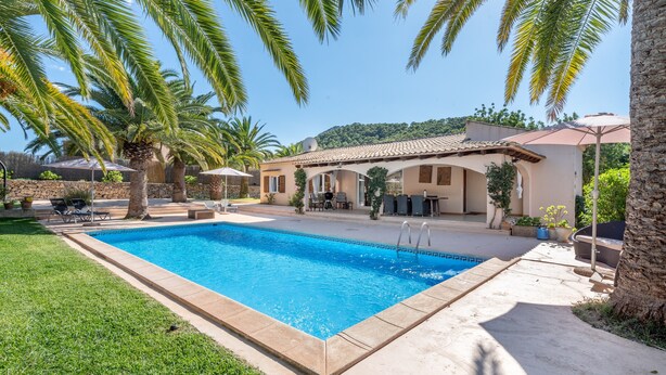 A swimming pool with a tiled deck, surrounded by palm trees and a building with a tiled roof. - Holiday Home “Polita des Carritxo” with Mountain View, Pool, Garden & Terrace (Es Carritxo)