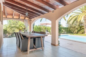 An outdoor patio with a wooden table and chairs, a hanging light fixture, and a view of a swimming pool and greenery.