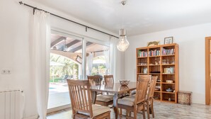 A dining area with a wooden table and chairs, a bookshelf, and a view through a glass door to an outdoor patio. - Holiday Home “Polita des Carritxo” with Mountain View, Pool, Garden & Terrace (Es Carritxo)