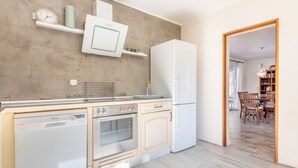 A kitchen with a dishwasher, oven, and refrigerator, wooden cabinets, a microwave, and a dining area with a table and chairs. - Holiday Home “Polita des Carritxo” with Mountain View, Pool, Garden & Terrace (Es Carritxo)