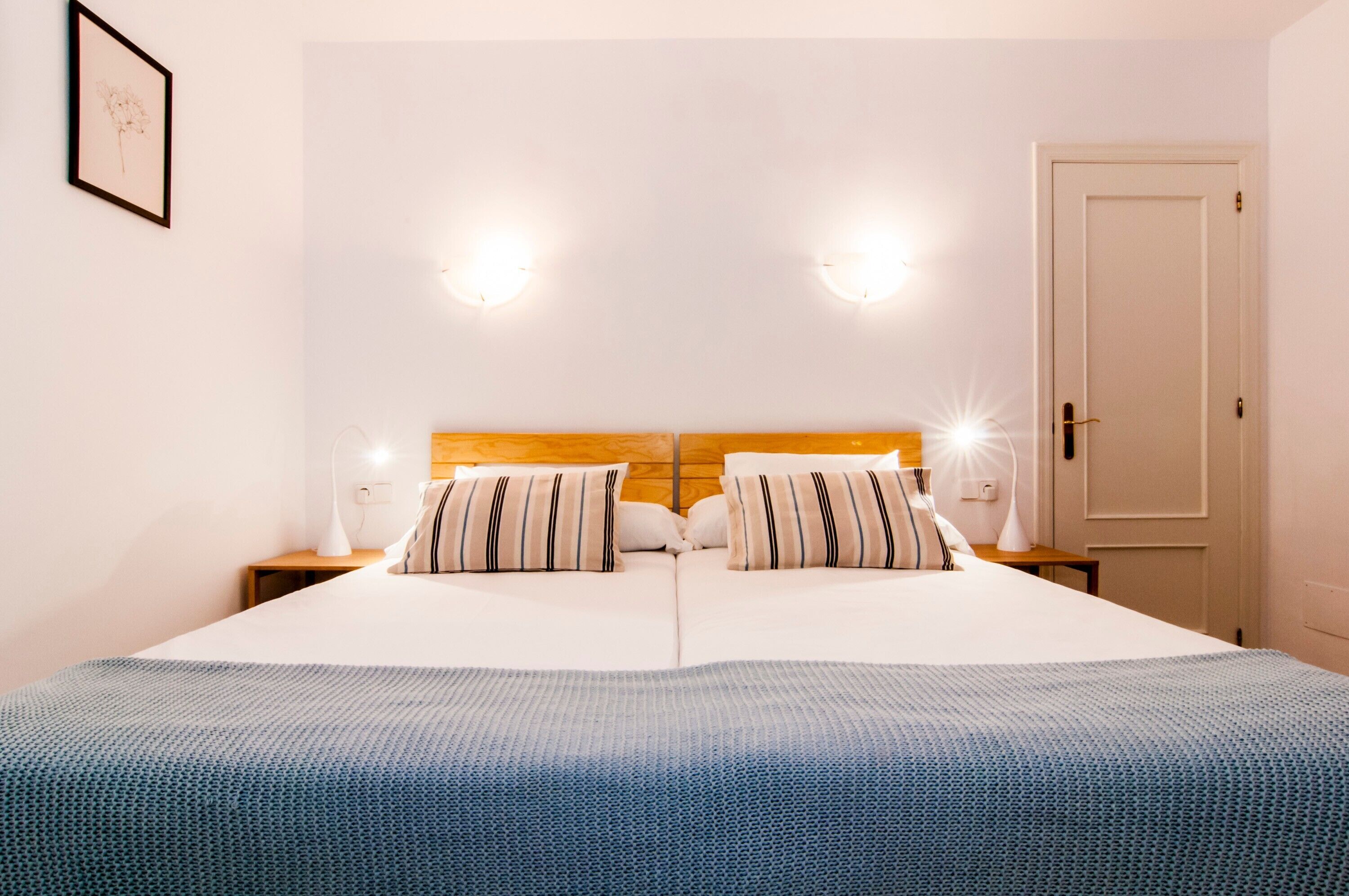 Un lit double avec une tête de lit en bois, deux tables de chevet avec des lampes, un tableau encadré au mur et une porte sur la droite.