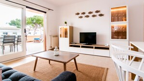 A modern living room with a TV, white furniture, a glass door leading outside, and decorative wall art. - Apartment “Drac Blanc 1-3” with Sea View, Balcony, Garden & Air Conditioning (Cala Ratjada)
