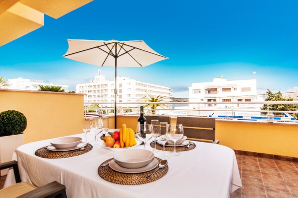 A dining table set with plates, bowls, wine glasses, and a fruit bowl, with a view of buildings in the background.