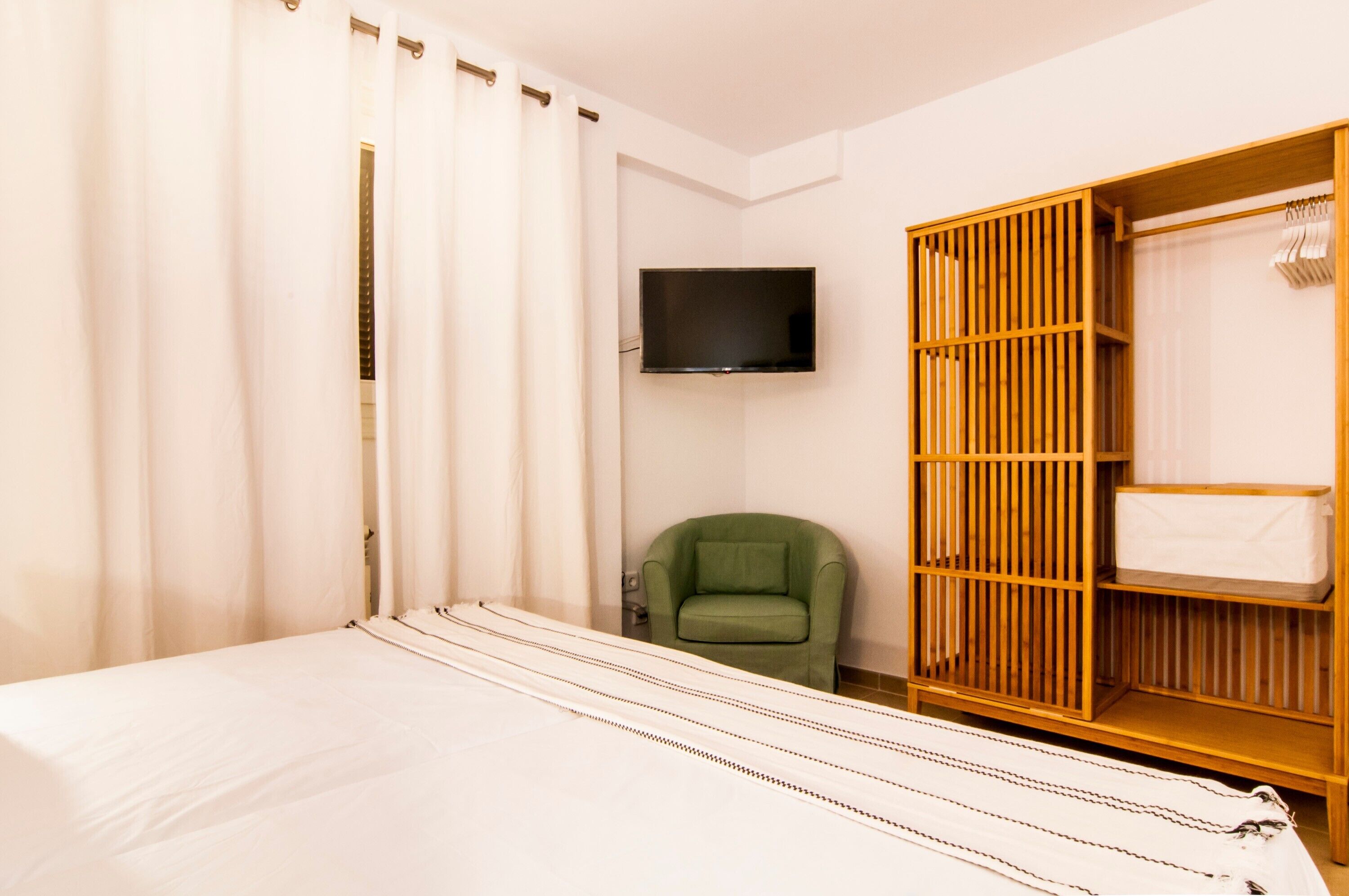 Une chambre d’hôtel avec un lit, un fauteuil vert, un téléviseur fixé au mur et une armoire en bois avec un miroir.